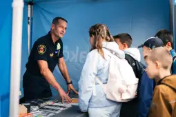 Mężczyzna w mundurze rozmawia z grupą dzieci przy stoisku informacyjnym.