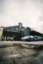 Autobus stojący przed budynkiem, dzieci w kamizelkach odblaskowych czekają w kolejce.