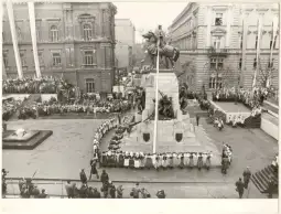 Uroczystość z udziałem tłumu ludzi wokół pomnika, flagi i budynków w tle.