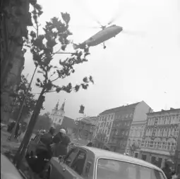 Helikopter transportujący ładunek nad miastem, w tle budynki i grupa ludzi.