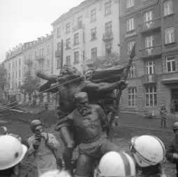 Pomnik przedstawiający postacie historyczne w miejskim otoczeniu, otoczony przez ludzi i policję.