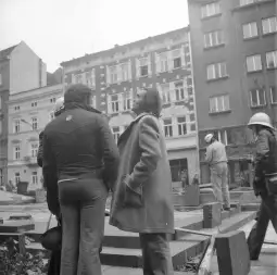 Grupa ludzi na placu budowy, w tle budynki i prace remontowe.
