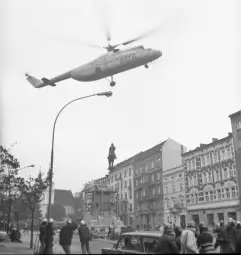 Helikopter transportujący pomnik w miejskim krajobrazie z ludźmi obserwującymi sytuację.