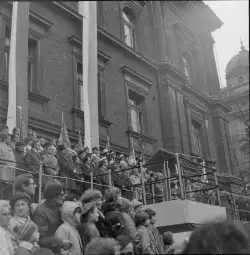 Tłum ludzi zgromadzony przed budynkiem, z flagami i przemawiającymi na podium.