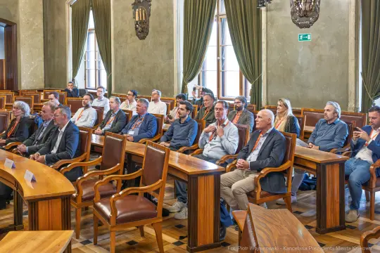 Grupa ludzi siedzących w sali obrad, zwróconych w stronę mównicy.