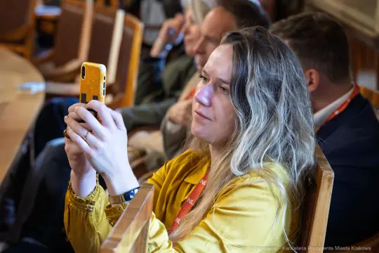 Kobieta z długimi włosami trzyma telefon i robi zdjęcie w sali konferencyjnej.