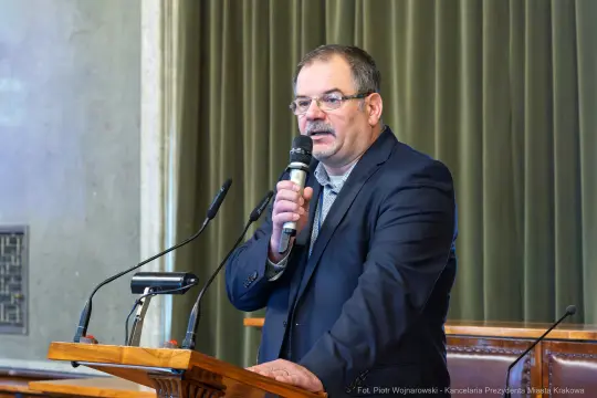 Mężczyzna w garniturze przemawia do mikrofonu na tle zielonej zasłony.