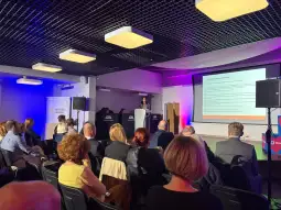 Konferencja z prelekcją, uczestnicy siedzą na sali, prezentacja na ekranie.