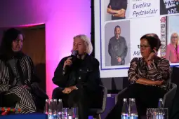 Panel dyskusyjny z trzema kobietami, jedna z mikrofonem, w tle prezentacja.