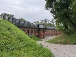 Forteca z cegły otoczona zielenią, z widocznymi schodami i tarasem.