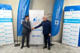 Dwaj mężczyźni uścisnęli sobie dłonie, w tle flagi i banery z logo Gulermak oraz napisami związanymi z Krakowem.