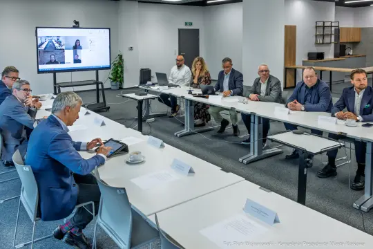 Spotkanie w sali konferencyjnej z uczestnikami przy stole i prezentacją na ekranie.