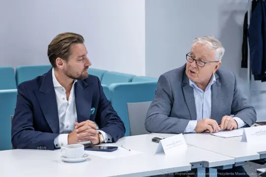 Dwóch mężczyzn rozmawia przy stole, jeden z nich trzyma notatki.