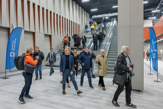 Grupa ludzi w nowoczesnym wnętrzu, schody ruchome w tle, niektórzy w odzieży roboczej.