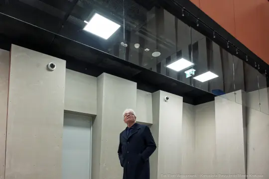 Mężczyzna w płaszczu stoi w nowoczesnym wnętrzu z betonowymi ścianami i sufitami.