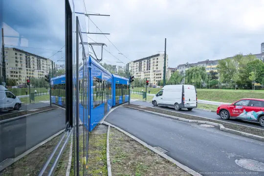 Tramwaj skręcający w lewo, obok przejeżdżający samochód dostawczy i czerwony pojazd.
