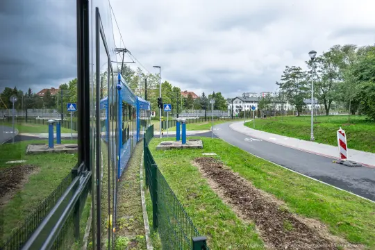 Widok na torowisko tramwajowe z zielonym otoczeniem i budynkami w tle.