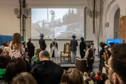 Widownia podczas występu teatralnego z historycznymi postaciami i projekcją w tle.