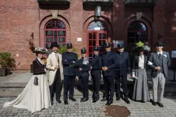 Grupa osób w strojach historycznych, pozująca przed ceglanym budynkiem.