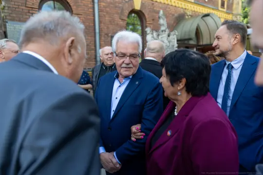 Grupa osób rozmawia na tle budynku, wśród nich mężczyzna w okularach i kobieta w fioletowej marynarce.