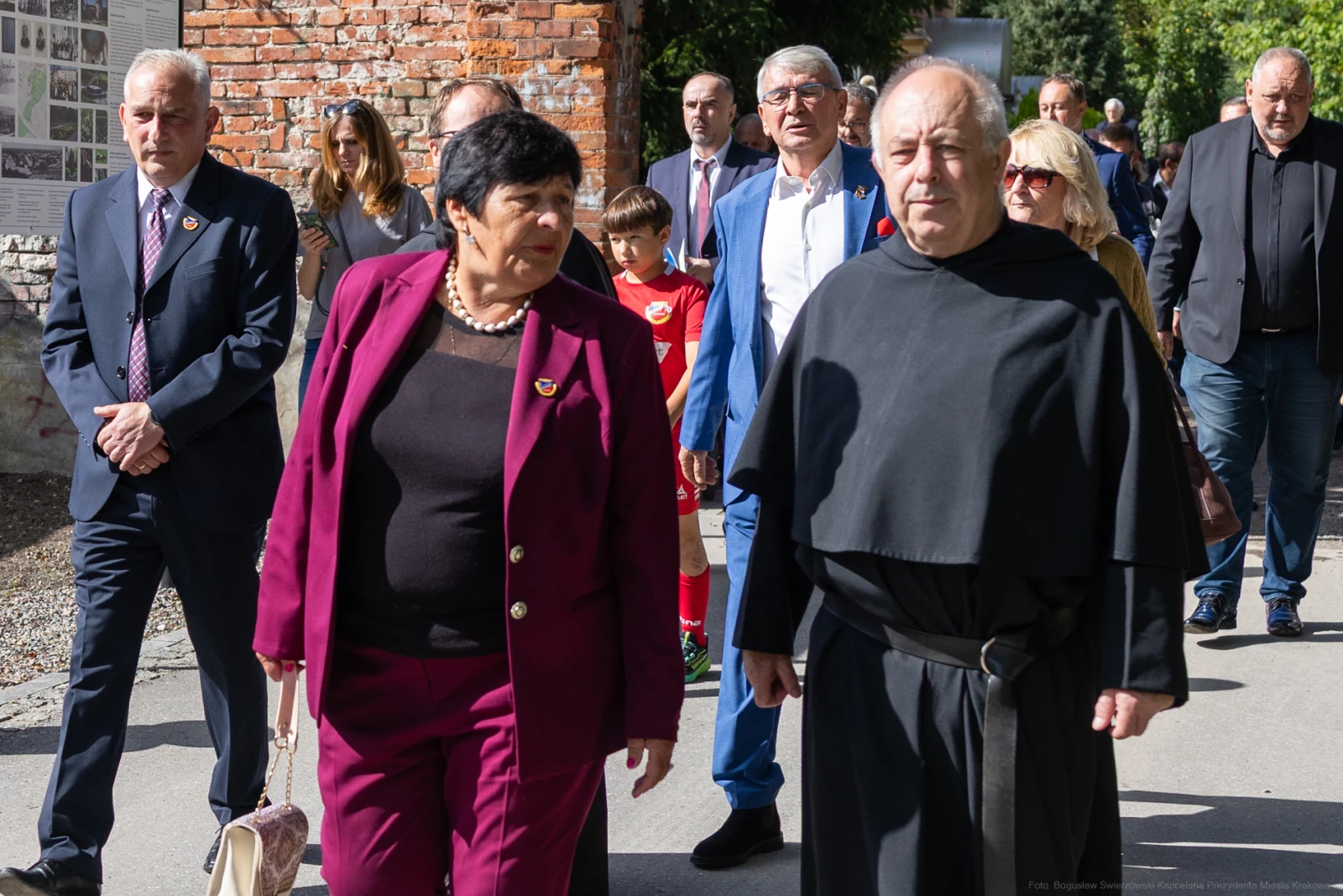 Grupa osób w formalnych strojach spaceruje po ulicy, wśród nich mężczyzna w habicie.  Autor: Bogusław Świerzowski