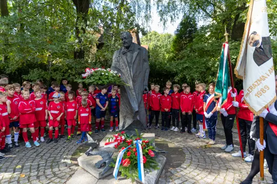 Grupa dzieci w strojach sportowych składa hołd przy pomniku, z kwiatami i flagami.