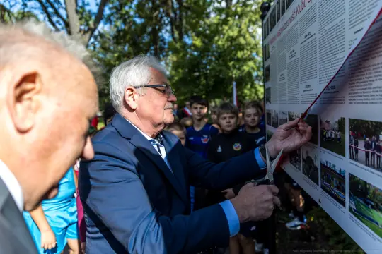 Mężczyzna przecina wstęgę przy tablicy informacyjnej, w tle grupa dzieci.