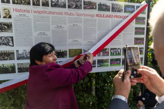 Kobieta przecina wstęgę przy tablicy upamiętniającej historię Klubu Sportowego Kolejarz-Prokocim.