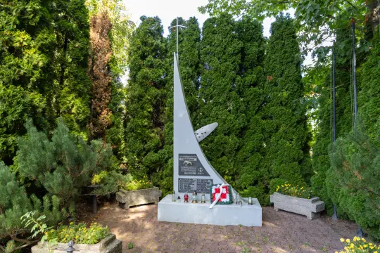 Pomnik upamiętniający wydarzenia historyczne, otoczony zielenią i kwiatami.