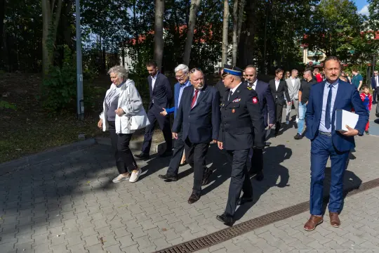 Grupa osób w garniturach i mundurach idzie alejką w parku.