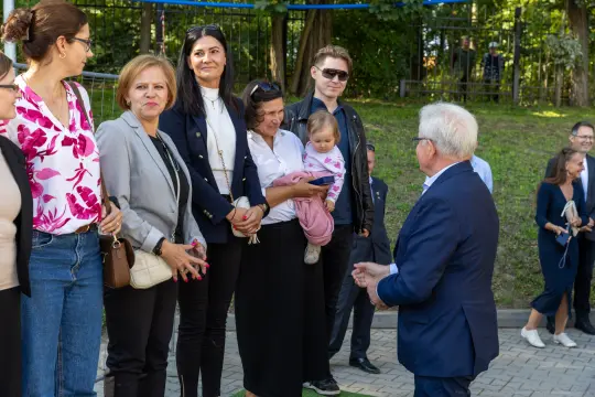 Grupa osób rozmawia w parku, jedna z kobiet trzyma dziecko na rękach.