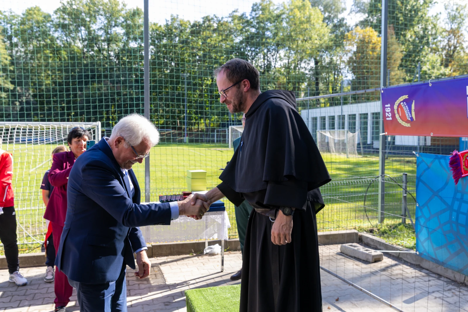 Mężczyzna w garniturze podaje rękę mnichowi w habicie na boisku sportowym.  Autor: Bogusław Świerzowski