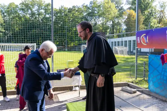 Mężczyzna w garniturze podaje rękę mnichowi w habicie na boisku sportowym.