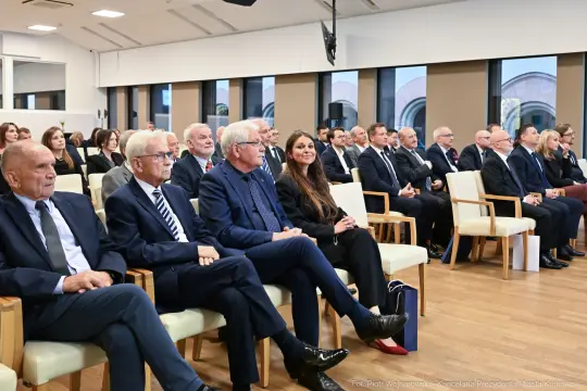 Grupa ludzi siedzących w sali konferencyjnej, ubranych w eleganckie stroje.
