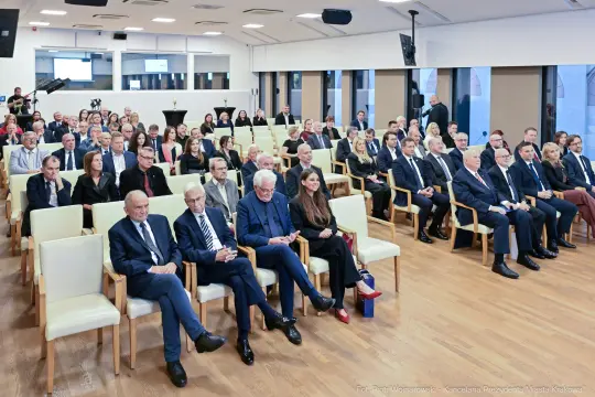 Zgromadzenie osób w sali konferencyjnej, siedzących w rzędach, w formalnych strojach.