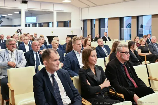 Grupa ludzi siedzących w sali konferencyjnej, uczestniczących w wydarzeniu.