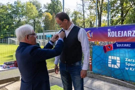 Mężczyzna w garniturze przypina odznaczenie do koszuli innego mężczyzny na tle boiska.