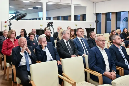 Grupa ludzi siedzących w sali konferencyjnej, niektórzy w garniturach, w trakcie wydarzenia.