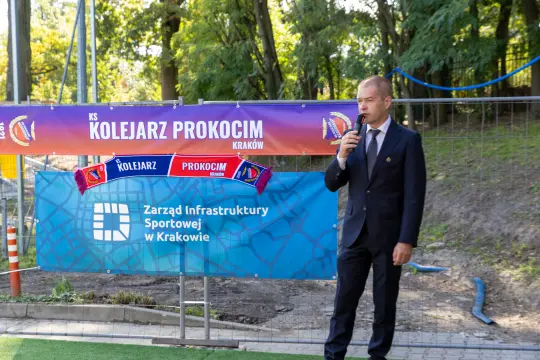 Mężczyzna w garniturze przemawia na tle bannerów związanych z klubem sportowym.
