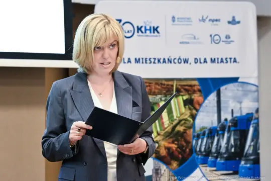 Kobieta w garniturze trzyma dokumenty, przemawia na tle banera promującego miasto.
