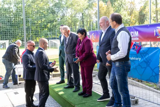 Uroczystość z udziałem kilku osób na zielonym dywanie, w tle boisko sportowe.