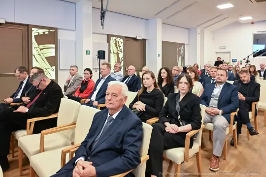 Grupa ludzi siedzących w sali konferencyjnej, niektórzy z telefonami, inni w rozmowach.