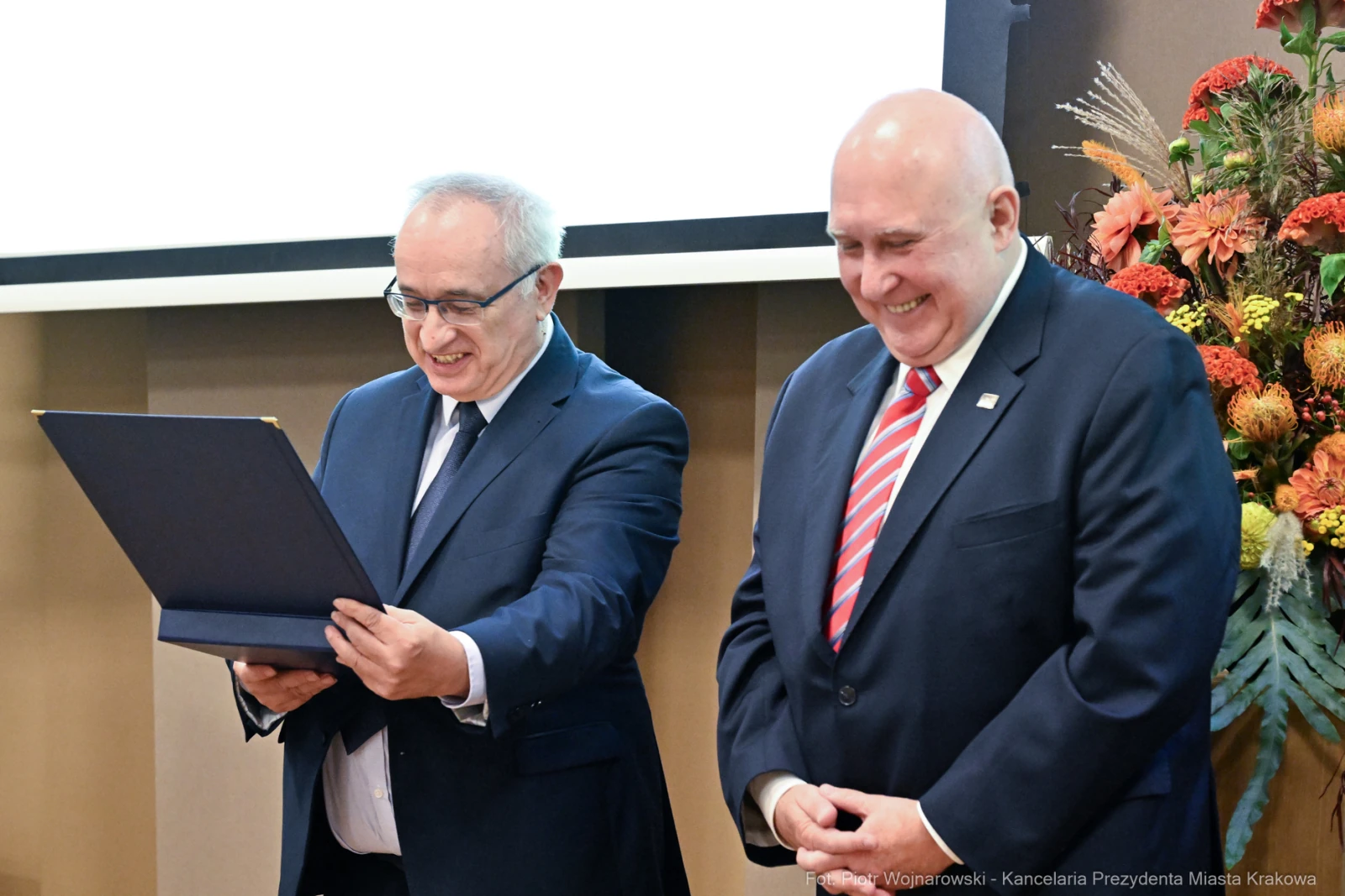Dwaj mężczyźni uśmiechają się, jeden trzyma dyplom, drugi go obserwuje.  Autor: Piotr Wojnarowski