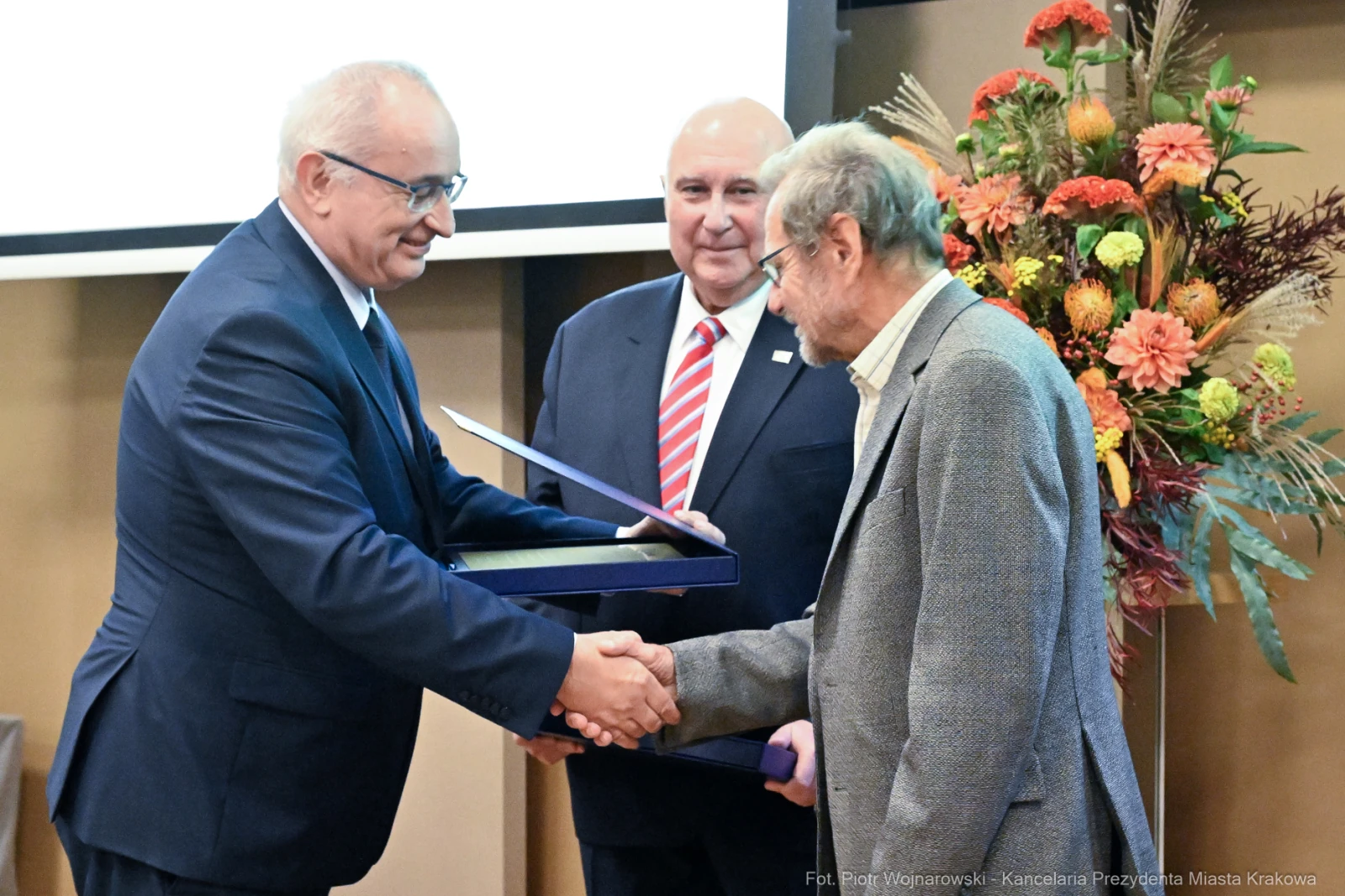 Wręczenie nagród, uściski dłoni, uśmiechnięci mężczyźni w formalnych strojach. Kwiaty w tle.  Autor: Piotr Wojnarowski