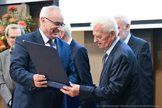 Wręczenie nagrody przez uśmiechniętego mężczyznę starszemu mężczyźnie w garniturze.