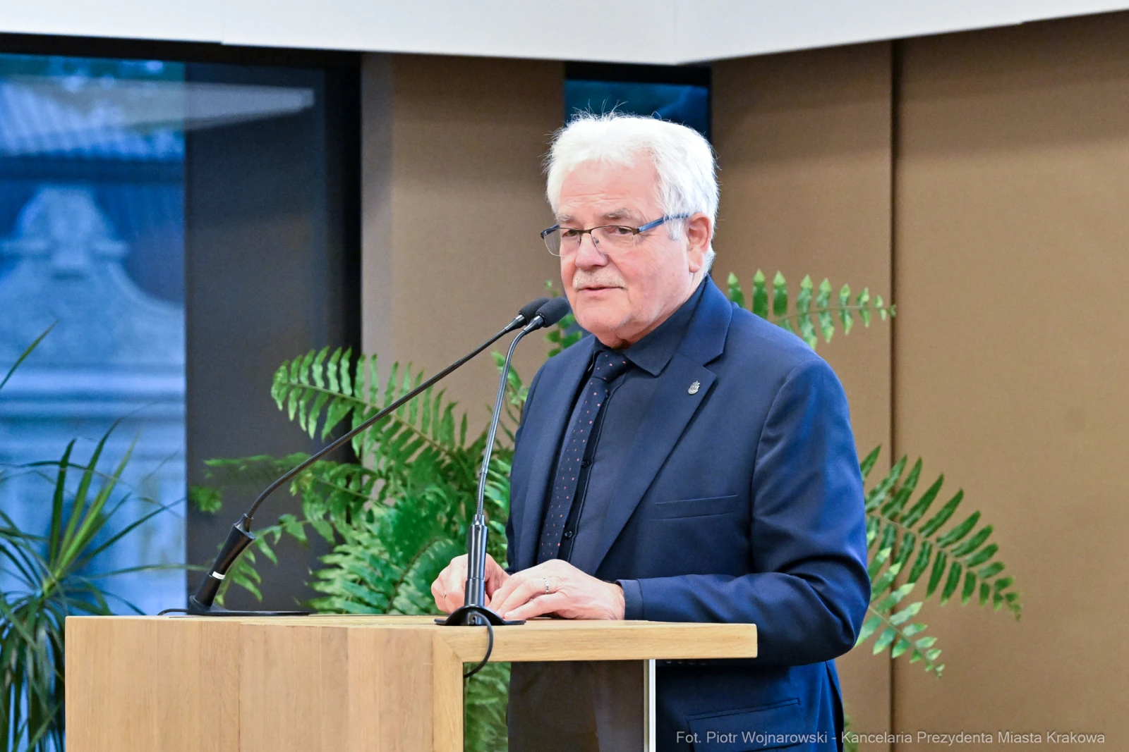Mężczyzna w garniturze przemawia przy mównicy, w tle rośliny i elementy architektury.  Autor: Piotr Wojnarowski