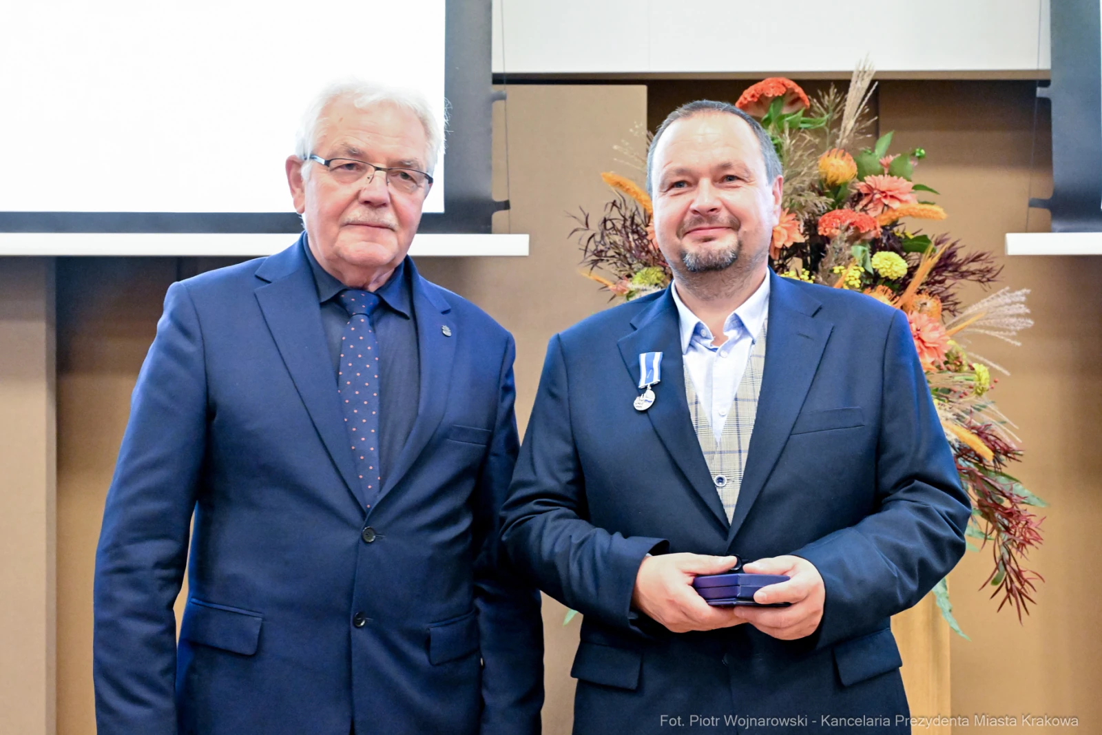 Dwaj mężczyźni w garniturach, jeden z medalem, stoją obok siebie na tle dekoracji.  Autor: Piotr Wojnarowski