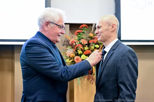 Mężczyzna w garniturze odbiera odznaczenie od innego mężczyzny w eleganckim stroju.