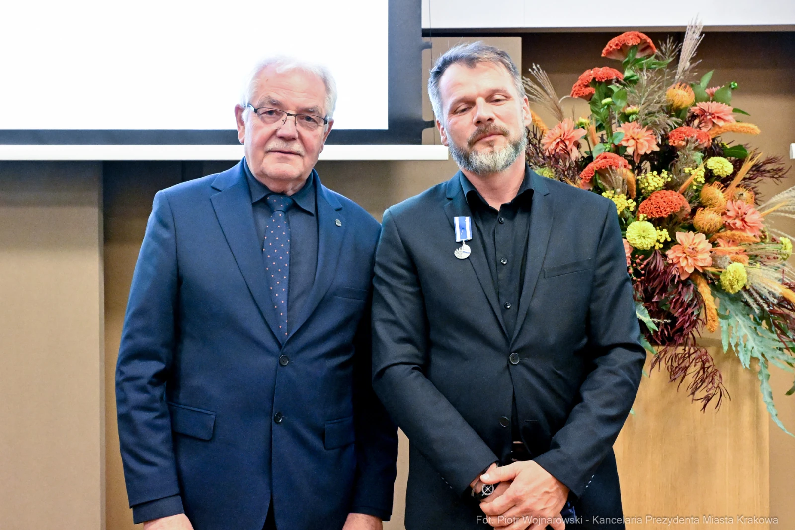 Dwaj mężczyźni w formalnych strojach, jeden z medalem, w tle dekoracje kwiatowe.  Autor: Piotr Wojnarowski