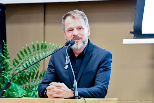 Mężczyzna w garniturze przemawia do mikrofonu, w tle roślinność.